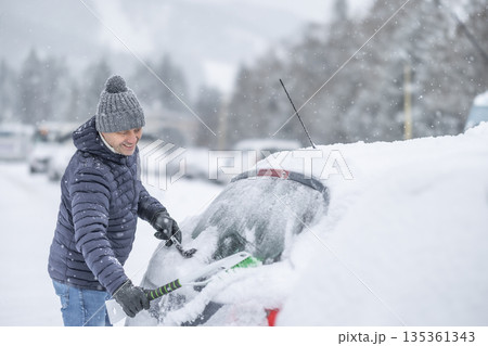 A man in a dark jacket and gray hat diligently removes a thick layer of fresh snow from the windshield of his car with a scraper and a broom. The entire scene takes place during a heavy snowfall in a 135361343