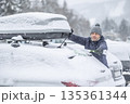 A man in a blue jacket and knit hat uses a broom to clear a thick layer of snow from the back of his car. The surrounding snowy landscape and the roof box on the vehicle suggest that he is preparing 135361344
