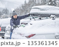 A man in a blue jacket and knit hat uses a broom to clear a thick layer of snow from the back of his car. The surrounding snowy landscape and the roof box on the vehicle suggest that he is preparing 135361345