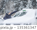 A man in a dark jacket and gray hat diligently removes a thick layer of fresh snow from the windshield of his car with a scraper and a broom. The entire scene takes place during a heavy snowfall in a 135361347