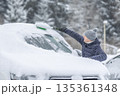 A man in a dark jacket and gray hat diligently removes a thick layer of fresh snow from the windshield of his car with a scraper and a broom. The entire scene takes place during a heavy snowfall in a 135361348