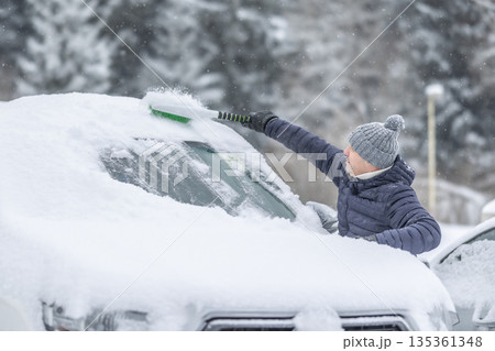A man in a dark jacket and gray hat diligently removes a thick layer of fresh snow from the windshield of his car with a scraper and a broom. The entire scene takes place during a heavy snowfall in a 135361348