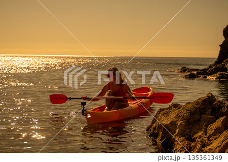 Woman, kayak, ocean. Woman paddles an orange kayak on sparkling water near rocky coastline at sunset with copy space. 135361349