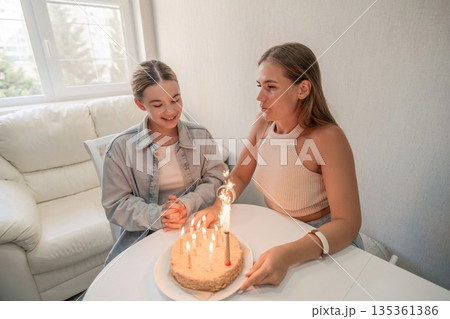 Birthday Cake Mother Daughter Celebration: Woman blows out candles on birthday cake with daughter at home joyful family moment. 135361386