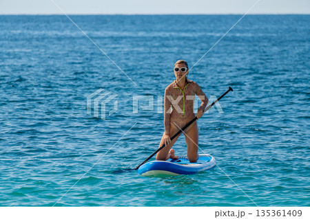 Woman, paddleboard, ocean. Active female kneeling on a stand up paddleboard in clear blue water enjoying summer sports with copy space. 135361409