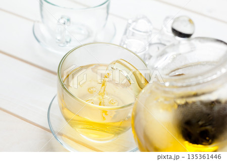 Hot tea is poured into a glass while ice cubes are placed nearby on a table 135361446