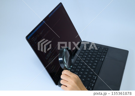 虫眼鏡でPCのプログラムコードを確認する手元 135361878