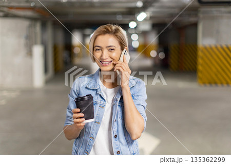 A smiling woman in a denim jacket holds a coffee cup while talking on her phone in a parking garage A smiling woman in a denim jacket holds a coffee cup while talking on her phone in a parking garage 135362299