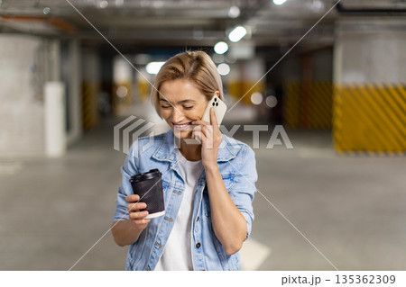 A smiling woman holds a coffee cup while talking on her smartphone in a parking garage A smiling woman holds a coffee cup while talking on her smartphone in a parking garage 135362309