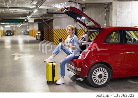 A woman sits on the trunk of a red car in a parking garage, enjoying coffee and using a tablet 135362322