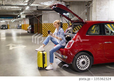 A woman sits in the open trunk of a red car in an underground parking garage, using her phone and enjoying a coffee 135362324