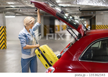 A woman smiles while loading a yellow suitcase into the trunk of a red car in a parking garage 135362343