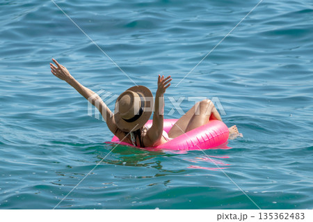 Woman Floating Sea: Woman relaxing on pink inflatable in blue sea, hat on, arms raised, summer fun. Woman Floating Sea: Woman relaxing on pink inflatable in blue sea, hat on, arms raised, summer fun. 135362483