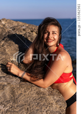 Woman, beach, rocks. Happy young woman smiling, leaning on sunlit rocks by the ocean with copy space. Woman, beach, rocks. Happy young woman smiling, leaning on sunlit rocks by the ocean with copy space. 135362561
