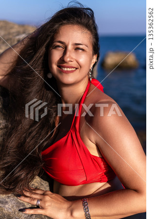 Woman, beach, smile, beautiful woman with long dark hair posing happily in a red bikini top by the sea on a sunny day. Woman, beach, smile, beautiful woman with long dark hair posing happily in a red bikini top by the sea on a sunny day. 135362562