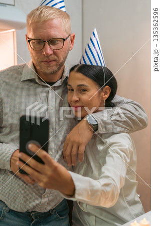 Couple Selfie Birthday Party Home: Smiling couple wearing party hats takes selfie indoors, celebrating birthday. Couple Selfie Birthday Party Home: Smiling couple wearing party hats takes selfie indoors, celebrating birthday. 135362596