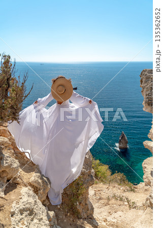 Woman, sea, cliff. Woman in flowing white dress, straw hat, stands on cliff overlooking vast blue sea. Summer travel freedom. Woman, sea, cliff. Woman in flowing white dress, straw hat, stands on cliff overlooking vast blue sea. Summer travel freedom. 135362652
