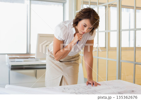 Woman wearing white blouse leaning over blueprints on table in office reviewing architectural plans Woman wearing white blouse leaning over blueprints on table in office reviewing architectural plans 135362676