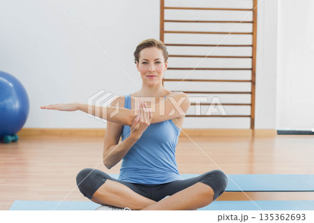 Woman stretching right arm across chest while holding left wrist on blue mat in exercise studio 135362693