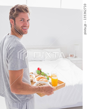 Man standing in bright bedroom holding wooden tray with cereal, croissant and red rose, copy space Man standing in bright bedroom holding wooden tray with cereal, croissant and red rose, copy space 135362710