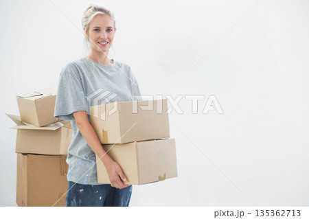 Woman holding two cardboard boxes while wearing paint-splattered T-shirt in white room, copy space 135362713