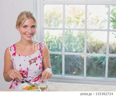 Woman in floral dress cutting salad using fork, knife at kitchen table by window, copy space 135362714