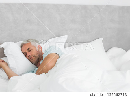 Senior male sleeping on side in bedroom with white duvet and pillows on grey upholstered headboard Senior male sleeping on side in bedroom with white duvet and pillows on grey upholstered headboard 135362718