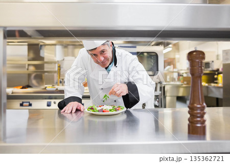 Male chef in uniform garnishing salad with herb sprig on stainless counter with pepper mill, ovens Male chef in uniform garnishing salad with herb sprig on stainless counter with pepper mill, ovens 135362721