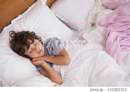 Boy sleeping on side in bed with white bedding, pink blanket and wooden headboard 135362723