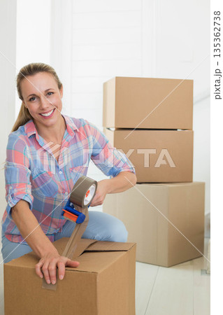 Mid adult woman kneeling on wood floor in entrance area sealing box with handheld tape dispenser Mid adult woman kneeling on wood floor in entrance area sealing box with handheld tape dispenser 135362738