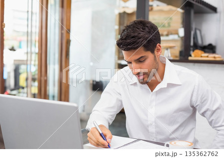 Man writing in notebook at café table beside laptop computer and coffee cup, copy space Man writing in notebook at café table beside laptop computer and coffee cup, copy space 135362762