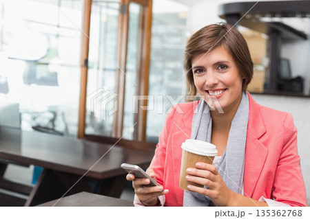 Woman wearing coral blazer holding smartphone and to-go coffee cup at coffee shop table, copy space Woman wearing coral blazer holding smartphone and to-go coffee cup at coffee shop table, copy space 135362768