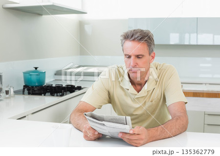 Man sitting and reading newspaper in kitchen at white countertop near turquoise cooking pot and mug Man sitting and reading newspaper in kitchen at white countertop near turquoise cooking pot and mug 135362779
