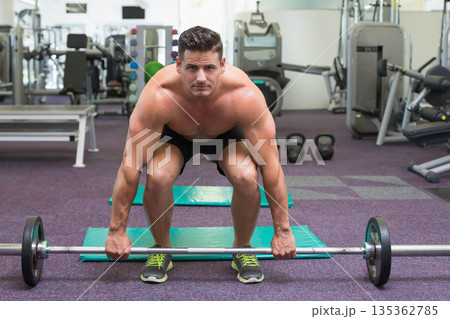 Shirtless man wearing black shorts lifting barbell on mat in gym with kettlebells and mirrored wall Shirtless man wearing black shorts lifting barbell on mat in gym with kettlebells and mirrored wall 135362785