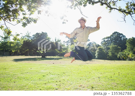 Man jumping barefoot on grassy lawn in park wearing straw fedora hat under dappled sunlight Man jumping barefoot on grassy lawn in park wearing straw fedora hat under dappled sunlight 135362788