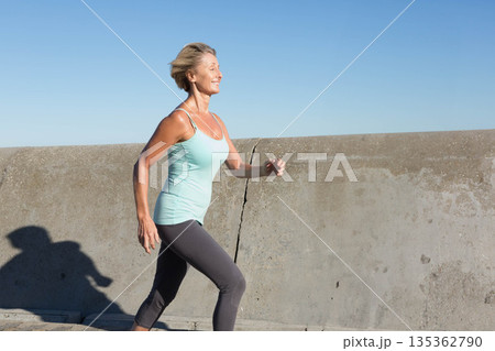Senior woman jogging on walkway by concrete sea wall in sportswear with athletic shoes, copy space 135362790