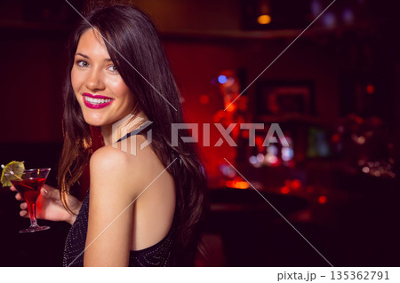 Woman wearing black dress standing by dimly lit bar counter holding red cocktail glass, copy space 135362791