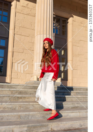 Fashion Woman Outfit Red Beret: Model poses on sunny day, wears red beret & sweater, white skirt, outside building. 135363157