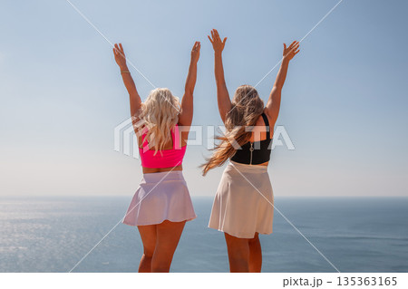 Two women are standing on a beach, one wearing a pink top and the other wearing a black top. They are both smiling and raising their arms in the air, as if they are celebrating something. 135363165