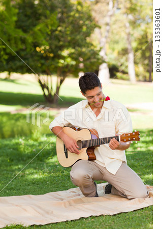 Mid adult man kneeling on picnic blanket in park, strumming guitar holding red rose between teeth Mid adult man kneeling on picnic blanket in park, strumming guitar holding red rose between teeth 135363601