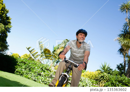 Mid-adult man riding bicycle wearing striped polo shirt and helmet along tropical garden pathway Mid-adult man riding bicycle wearing striped polo shirt and helmet along tropical garden pathway 135363602