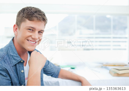 Man leaning, smiling toward camera at study desk by large window with books papers, copy space 135363613