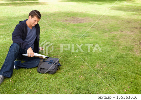 Male student sitting on grassy lawn flipping through book pages beside gray backpack, copy space 135363616