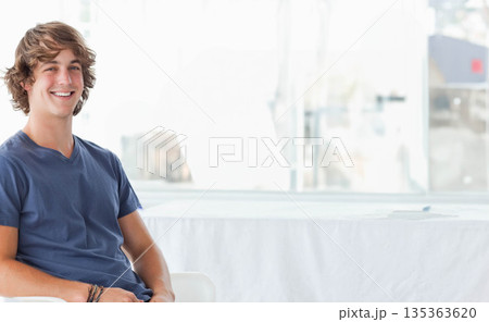 Man wearing V-neck shirt sitting on chair in lounge by table and large windows, copy space Man wearing V-neck shirt sitting on chair in lounge by table and large windows, copy space 135363620