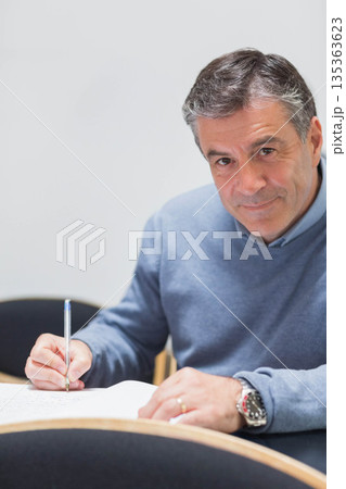 Middle-aged man writing in open notebook on wooden table in meeting room using silver pen Middle-aged man writing in open notebook on wooden table in meeting room using silver pen 135363623