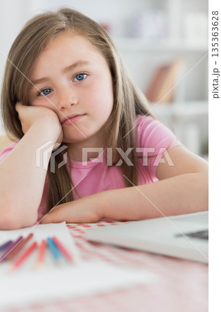 School-age girl resting cheek on hand at home study desk with colored pencils, paper and laptop 135363628