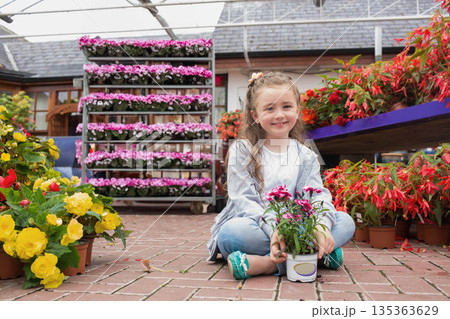 Female child sitting cross-legged holding white pot with pink blossoms on brick floor in greenhouse Female child sitting cross-legged holding white pot with pink blossoms on brick floor in greenhouse 135363629