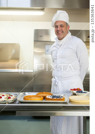 Male pastry chef standing behind steel prep table arranging fruit tarts in kitchen, copy space Male pastry chef standing behind steel prep table arranging fruit tarts in kitchen, copy space 135363641