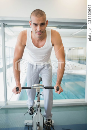 Man pedaling bike on mat gripping handlebars wearing tank top and sweatpants at fitness center Man pedaling bike on mat gripping handlebars wearing tank top and sweatpants at fitness center 135363665