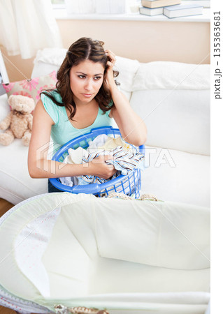 Woman sitting on white sofa holding blue laundry basket and scratching head beside baby bassinet Woman sitting on white sofa holding blue laundry basket and scratching head beside baby bassinet 135363681